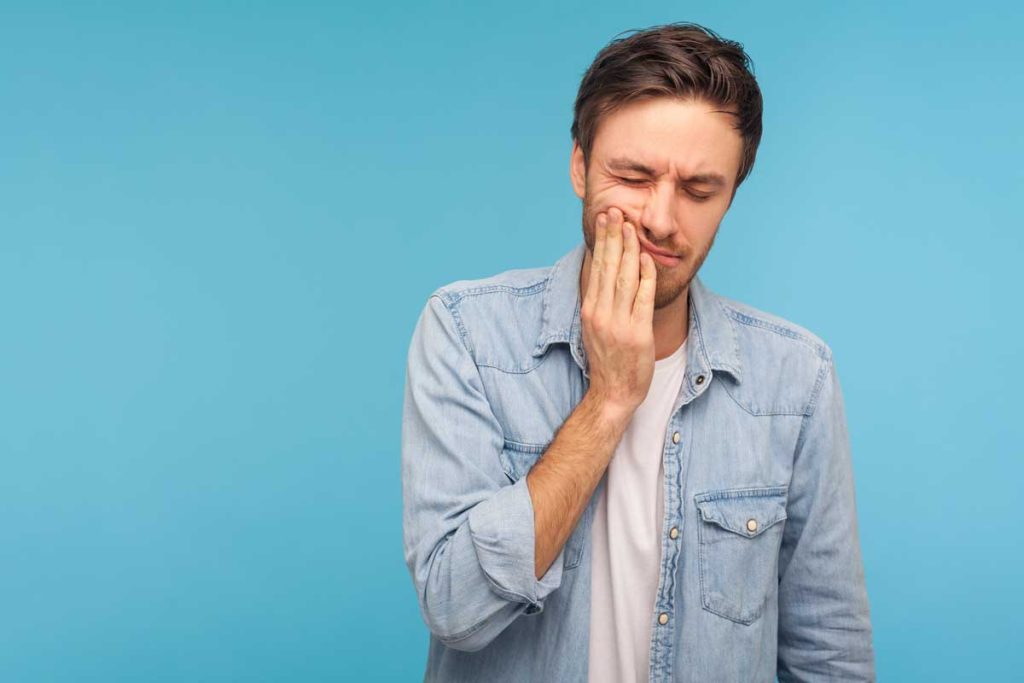 Dental problems. Portrait of man in denim shirt pressing sore cheek, suffering acute toothache, periodontal disease, cavities or jaw pain. indoor studio shot isolated on blue background.